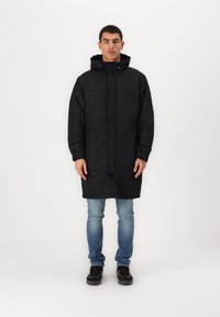 Svart vanntett parka med hette, med glidelås foran, to brystlommer, lange ermer og knelang design. Båret over lyse jeans.