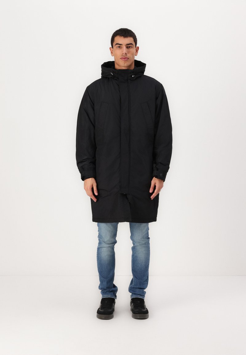 Svart vanntett parka med hette, med glidelås foran, to brystlommer, lange ermer og knelang design. Båret over lyse jeans.