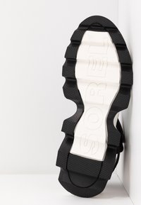 Semelle d'une chaussure de marque Sport noire et blanche avec des crampons profonds, positionnée verticalement contre un mur blanc d'angle.