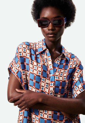 Vrouw met een donkere huid en afro kapsel draagt oversized rechthoekige zonnebril en een kort mouw overhemd met een blauw, wit en oranje patroon.