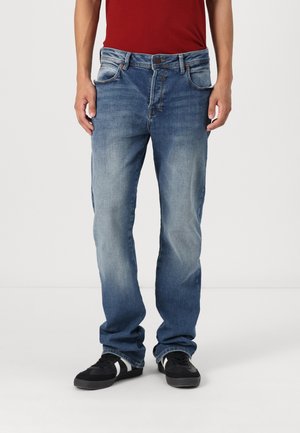 Jeans din denim, de culoare albastră deschisă până la medie, având un design cu picior drept, cinci buzunare și o ușoară decolorare. Purtați cu adidași negri.
