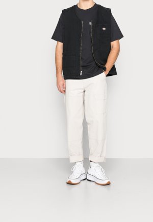 Mann trägt eine schwarze Zip-up-Weste über einem schwarzen T-Shirt, beige hochgekrempelte Cargo-Hose und weiße High-Top-Sneaker, steht mit einer Hand in der Tasche.