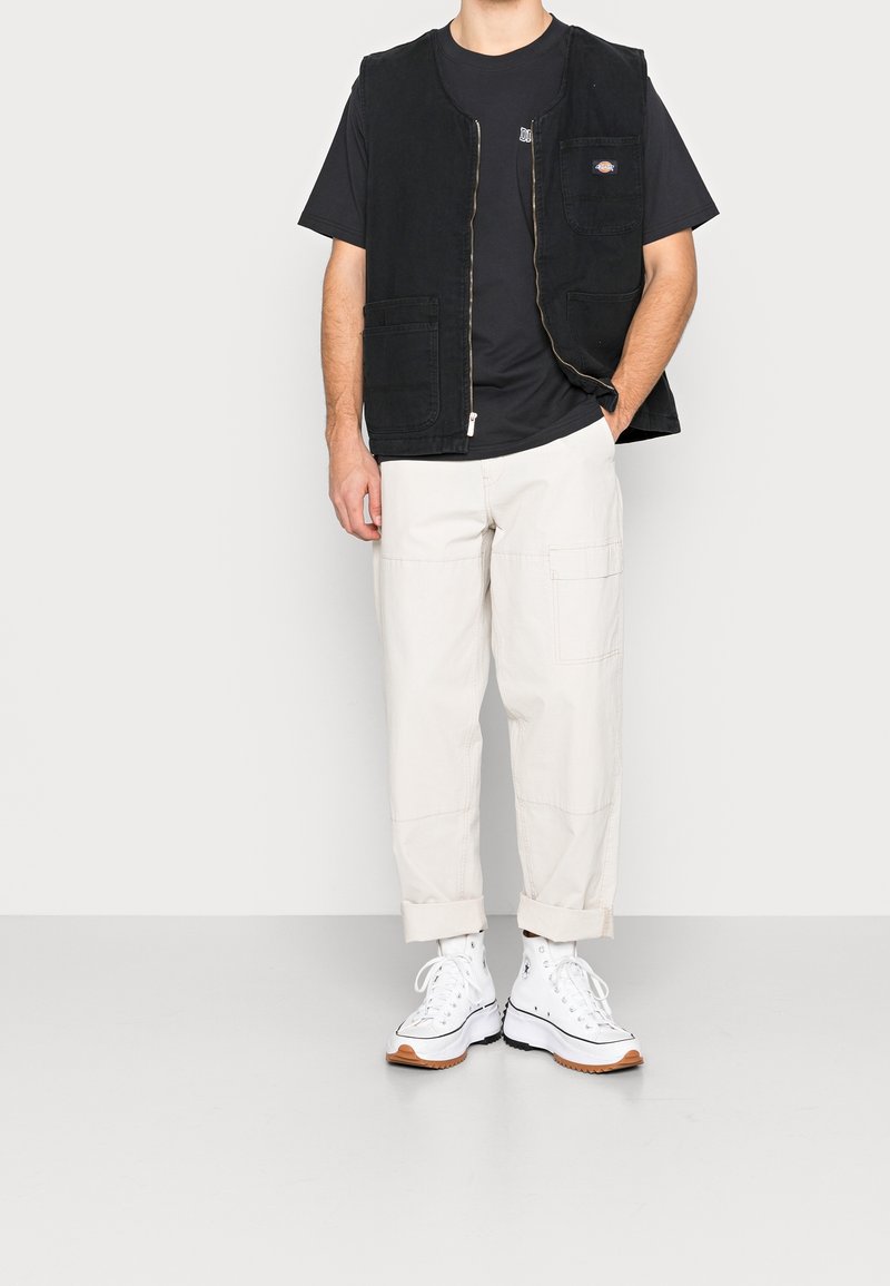 Homme portant un gilet zippé noir par-dessus un t-shirt noir, un pantalon cargo beige retroussé et des baskets montantes blanches, debout avec une main dans la poche.