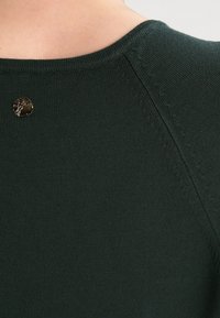 Ciemnozielony sweter dziany z okrągłym dekoltem, z małym złotym detalem na plecach. Gładka faktura z widocznymi szwami.