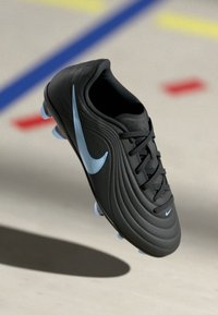 Μαύρο ποδοσφαιρικό παπούτσι με ελαφρώς μπλε σήμα Nike και τάπες, κρεμασμένο πάνω από μια λεία επιφάνεια με χρωματιστές γραμμές.