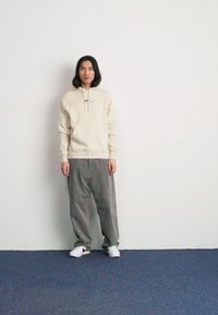 Beige hoodie med dragsnören, grå vida corduroybyxor och vita sneakers med lila detaljer. Modellen står mot en enkel vägg.