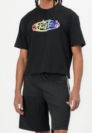 Osoba ubrana w czarny, skrócony t-shirt Adidas z tęczowym logo oraz czarne spodenki Adidas z białymi paskami i logo.