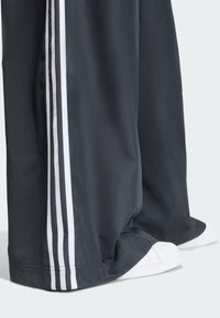 Pantalones deportivos negros de pierna ancha con franjas blancas en los laterales; combinados con zapatillas blancas. Tejido suave y ligero con un corte cómodo.