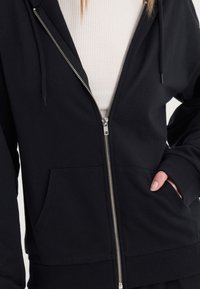 Schwarzer Kapuzenpullover zum Reißverschließen aus weichem Stoff, mit vorderem Reißverschluss, zwei seitlichen Taschen sowie einem gerippten Saum und Bündchen.