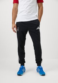 Sorte træningsbukser med hvide sidestriber, prydet med Tysklands nationale holds logo. Sammen med blå adidas sneakers og en hvid-rød skjorte.