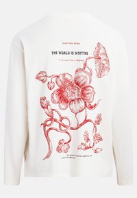 Camicia bianca a maniche lunghe con illustrazione botanica rossa e le frasi "REDEFINED REBEL", "THE WORLD IS WAITING" e testo in corsivo sul retro.