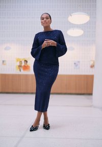 Granatowa, błyszcząca sukienka dwuczęściowa z długimi, rozszerzanymi rękawami i dopasowaną spódnicą. Połączona z czarnymi balerinami w białe kropki.