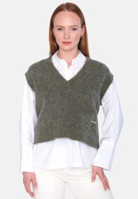 Grønn strikket vest med V-hals design, laget av et mykt, fluffy materiale, lagt over en hvit skjorte. Kjennetegnes av sin korte lengde.