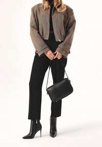 Nainen, jolla on päällään ruskea takki, mustat housut ja teräväkärkiset mustat korkokengät, pitää mustasta nahkakäsilaukusta varresta kiinni.