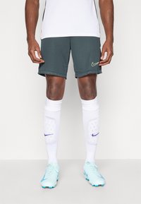 Nike nogometne kratke hlače u tamnozelenoj boji s žutim logom, uparene s bijelim štitnicima za noge i svijetloplavim kopačkama. Glatka tkanina, atletski kroj.