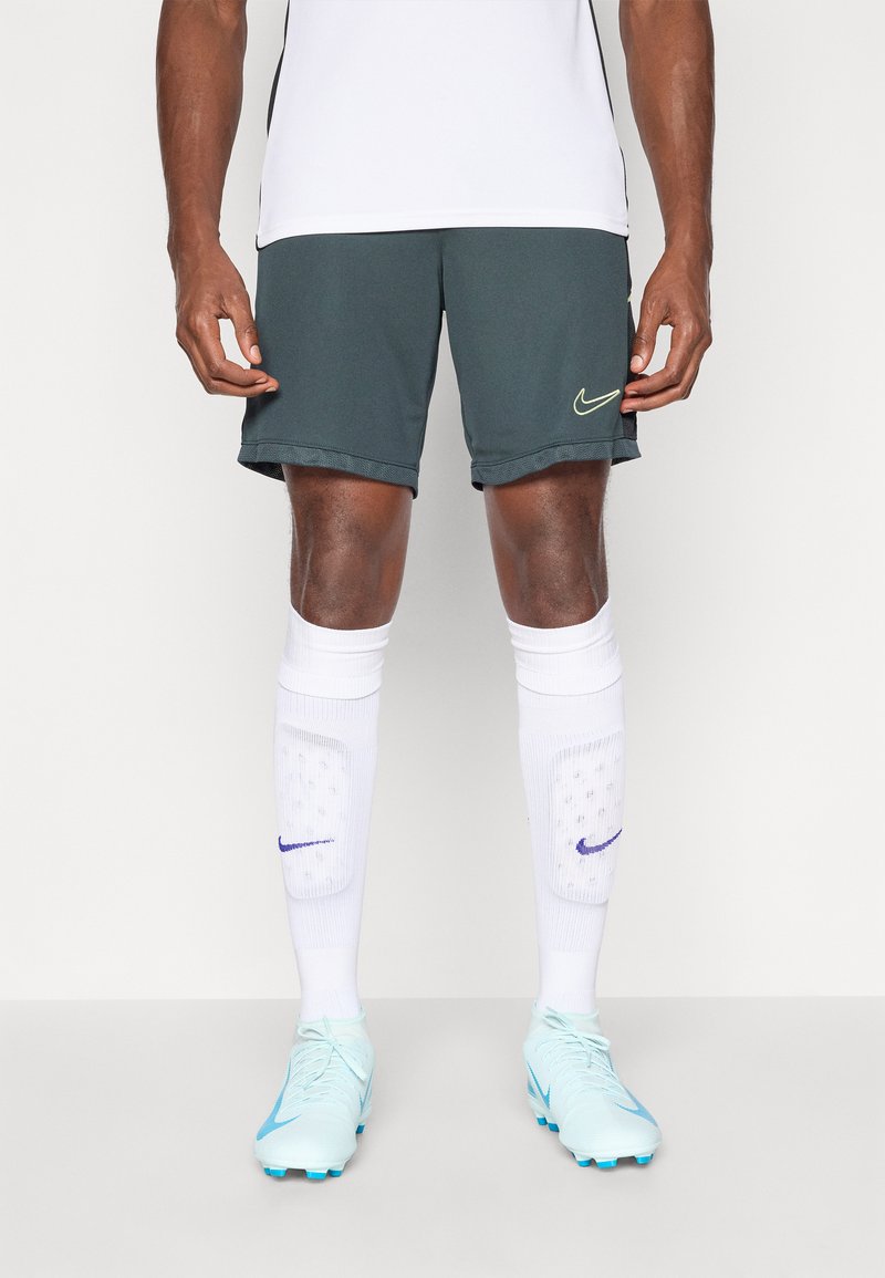 Nike nogometne kratke hlače u tamnozelenoj boji s žutim logom, uparene s bijelim štitnicima za noge i svijetloplavim kopačkama. Glatka tkanina, atletski kroj.