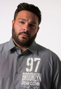 Camicia grigia con bottoni e colletto, decorata con il testo grafico bianco "97 BROOKLYN URBAN CLOTHING" sul lato sinistro. Texture liscia.