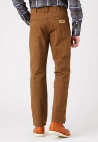 Man som bär bruna Wrangler-byxor, orange läderskor och en blå rutig långärmad skjorta, sedd bakifrån mot en vit bakgrund.