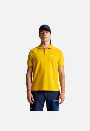 Man draagt een geel poloshirt met "SLAM"-logo, donkerblauwe broek met ritssluitingen en een zwarte pet, staand voor een effen achtergrond.