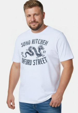 Uśmiechnięty mężczyzna z krótkimi włosami i brodą, ubrany w białą koszulkę z grafiką smoka i napisem "Soho Kitchen Oxford Street 2007" oraz niebieskie dżinsy.