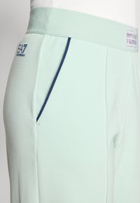 Calças de corrida em azul claro com cintura canelada, apresentando detalhes laterais em azul marinho e o logo EA7. Textura suave e corte ajustado.