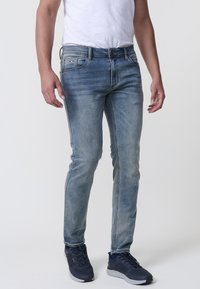 Karcsú szabású kék denim farmernadrág, elhalványult mosással, öt zsebbel és diszkrét varrási részletekkel. Tengerészkék sportcipővel viselve.