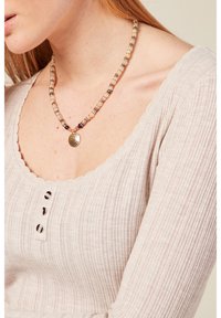 Die Perlenkette besteht aus hellen Steinen mit dunklen Akzenten und einem goldenen, strukturierten Anhänger. Sie wird mit einem gerippten, gewellten Oberteil in Beige getragen.