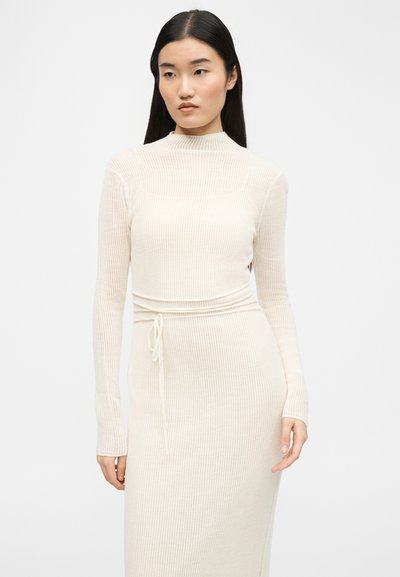 Femme aux longs cheveux noirs portant une robe en maille côtelée crème à manches longues avec une ceinture à la taille, se tenant devant un fond clair uni.