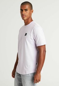 Jasnofioletowa bawełniana koszulka z krótkim rękawem, okrągłym dekoltem i małym czarnym logo na piersi. Noszona z wyblakłymi szarymi dżinsami.