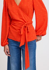 Top rouge à encolure en V profonde, avec des détails froncés sur les manches et une taille nouée. Texture à motif sur l'ensemble. Associé à un jean bleu à jambes larges.