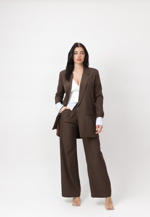 Kobieta stojąca, ubrana w brązowy, oversize'owy garnitur z szerokimi spodniami, białą bluzkę, mankiety w paski oraz przezroczyste sandały na otwartym palcu na jednolitym tle.