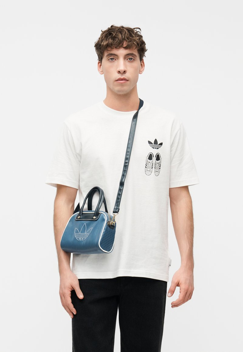 Giovane uomo che indossa una t-shirt bianca Adidas con logo e grafica di scarpa, che porta una piccola borsa Adidas blu con tracolla nera attraversata sul petto.