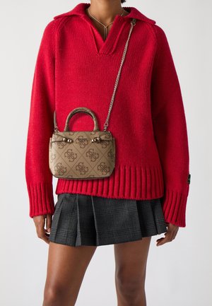 Femme portant un pull en tricot rouge et une jupe plissée à carreaux gris, portant un petit sac à main marron à motif avec une bandoulière en chaîne en travers du corps.