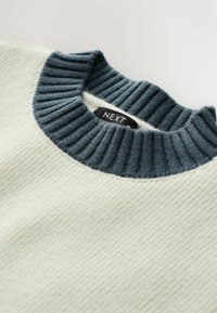 Pull crème avec un col côtelé bleu foncé. Présente un tissu texturé et une étiquette de marque visible à l'intérieur du col.