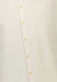 Camicia color crema realizzata in tessuto leggero, con chiusura frontale a bottoni composta da sei bottoni dorati e texture minimale.