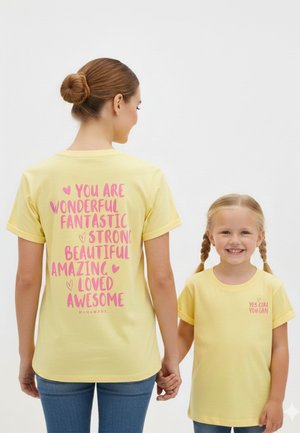 Frau und Mädchen halten sich an den Händen, tragen passende gelbe Shirts mit positivem pinkem Text; auf dem Rücken des Shirts der Frau stehen Affirmationen, auf der Vorderseite des Shirts des Mädchens steht "JA MÄDCHEN DU KANNST."