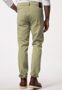 Pantalons en mélange de coton vert clair avec une coupe slim, présentant des poches arrière, des ourlets roulés et une étiquette noire au niveau de la ceinture. Bottes marron visibles.