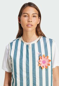 Striped sleeveless vest in blue and white with a zipper, featuring a pink flower patch and logo. Worn over a white t-shirt.