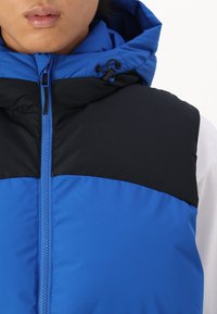 Gilet rembourré bleu et noir avec fermeture éclair, capuche réglable avec cordon, et une texture lisse. La partie supérieure présente un design en blocs de couleurs contrastantes.