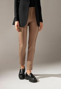 Pantaloni aderenti color cammello con una texture liscia e un tessuto elastico, abbinati a mocassini neri lucidi con una suola spessa.