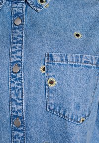 Camicia di jeans azzurro chiaro con colletto classico, diversi bottoni metallici e dettagli floreali ricamati sulla parte anteriore e sulla tasca.