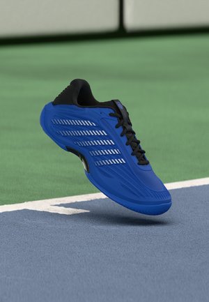 Blauwe sportschoen met zwarte veters zwevend boven een blauwgroen sportveld nabij een witte begrenzingslijn.