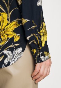 Main reposant le long du corps, portant une chemise bleu marine à imprimé floral jaune et blanc et un pantalon en cuir beige.