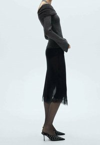 Zwarte off-the-shoulder gebreide top met lange mouwen, gecombineerd met een zwarte midi-rok met kantafwerking en een split, aangevuld met zwarte hoge muilen.