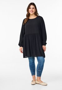 Svart långärmad tunika med en utsvängd nederdel, åtföljd av ljusblå denimleggings. Skorna är beige ballerinas med diskret rosett-detalj.