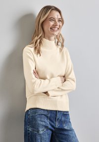 Maglione color crema con collo alto, texture a coste e orlo, abbinato a jeans blu, presentato su uno sfondo grigio chiaro.