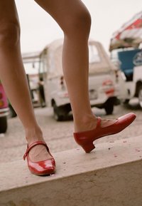 Czerwone lakierowane buty typu Mary Jane z niskim obcasem i paskiem wokół kostki, ustawione na teksturowanej powierzchni. W tle znajdują się miękkie, rozmyte pojazdy.