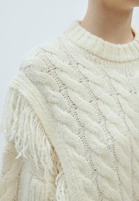 Chunky Creme Strickpullover mit Kabelmuster, geripptem Ausschnitt und Fransenverzierungen an den Schultern für zusätzlichen Textur.