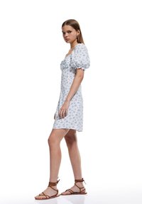 Vestido blanco con estampado floral azul, mangas abullonadas y escote cuadrado. Combinado con sandalias de tiras marrones. Textura de tela suave.