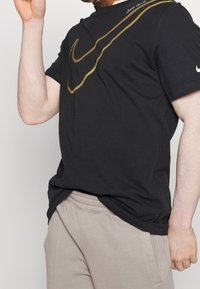 Osoba v černém tričku Nike s velkým zlatým swooshem a béžovými kalhotami, pózující s jednou rukou zvednutou u obličeje.